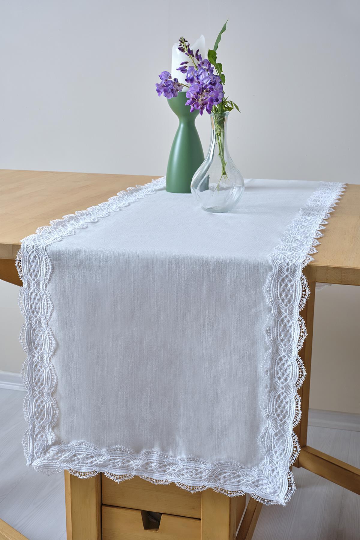 Weißer Tischläufer mit Spitzenkante, dekoriert mit einer Glasvase und lila Blumen auf einem Holztisch