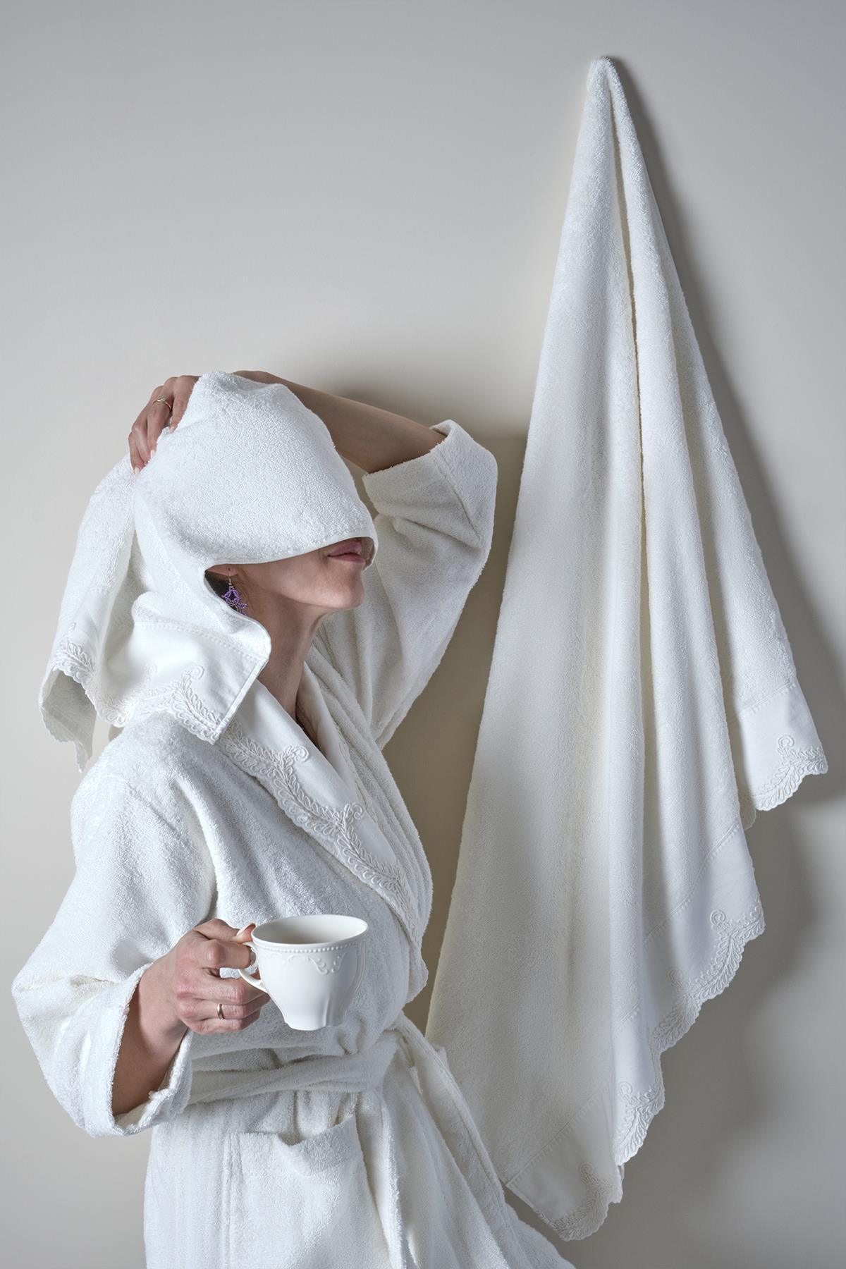 Frau im weißen Bambus-Bademantel hält eine Tasse in der Hand, im Hintergrund hängt ein Handtuch an der Wand