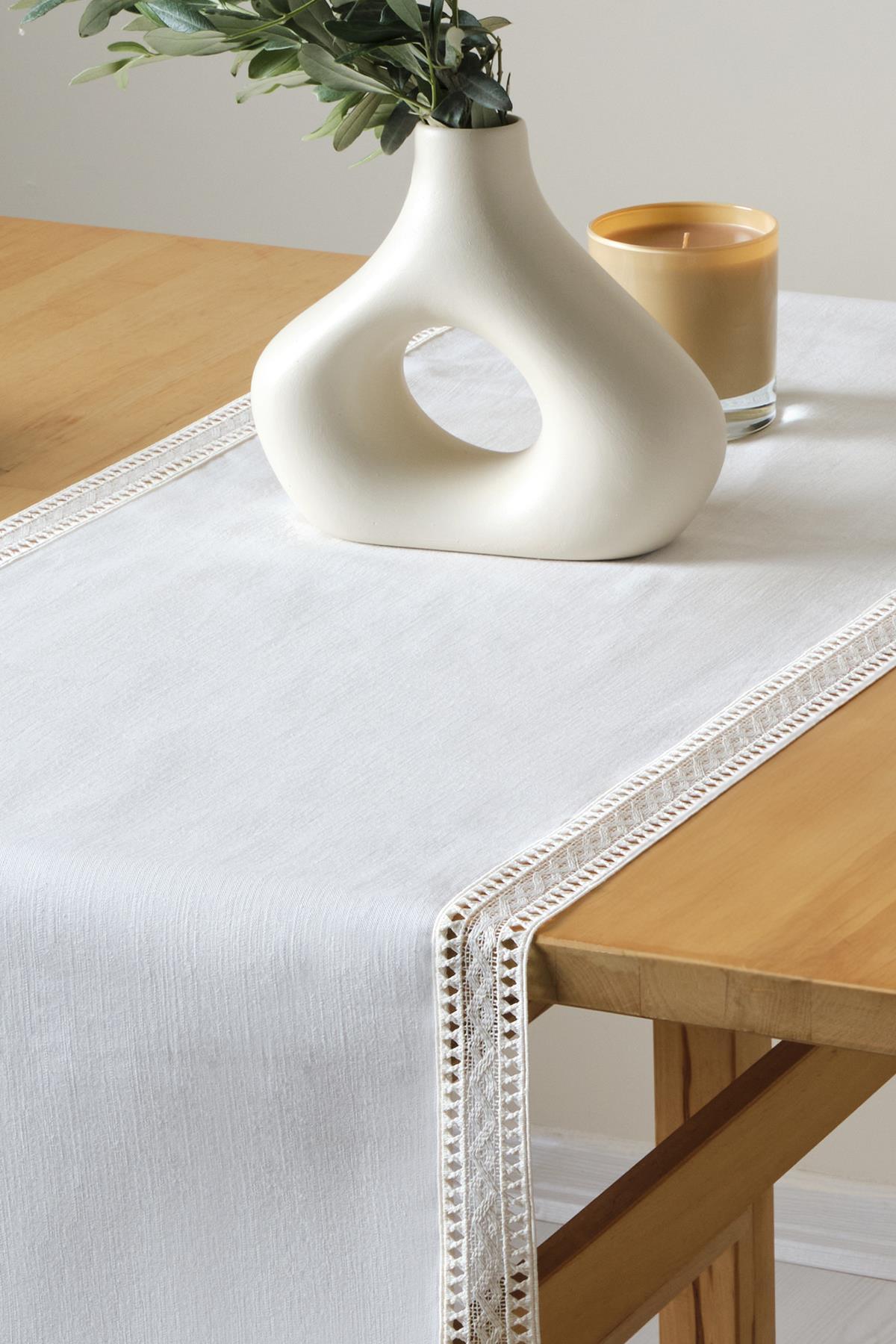 Dekorativer weißer Spitzenläufer auf einem Holztisch mit einer modernen Vase und einer Kerze.
