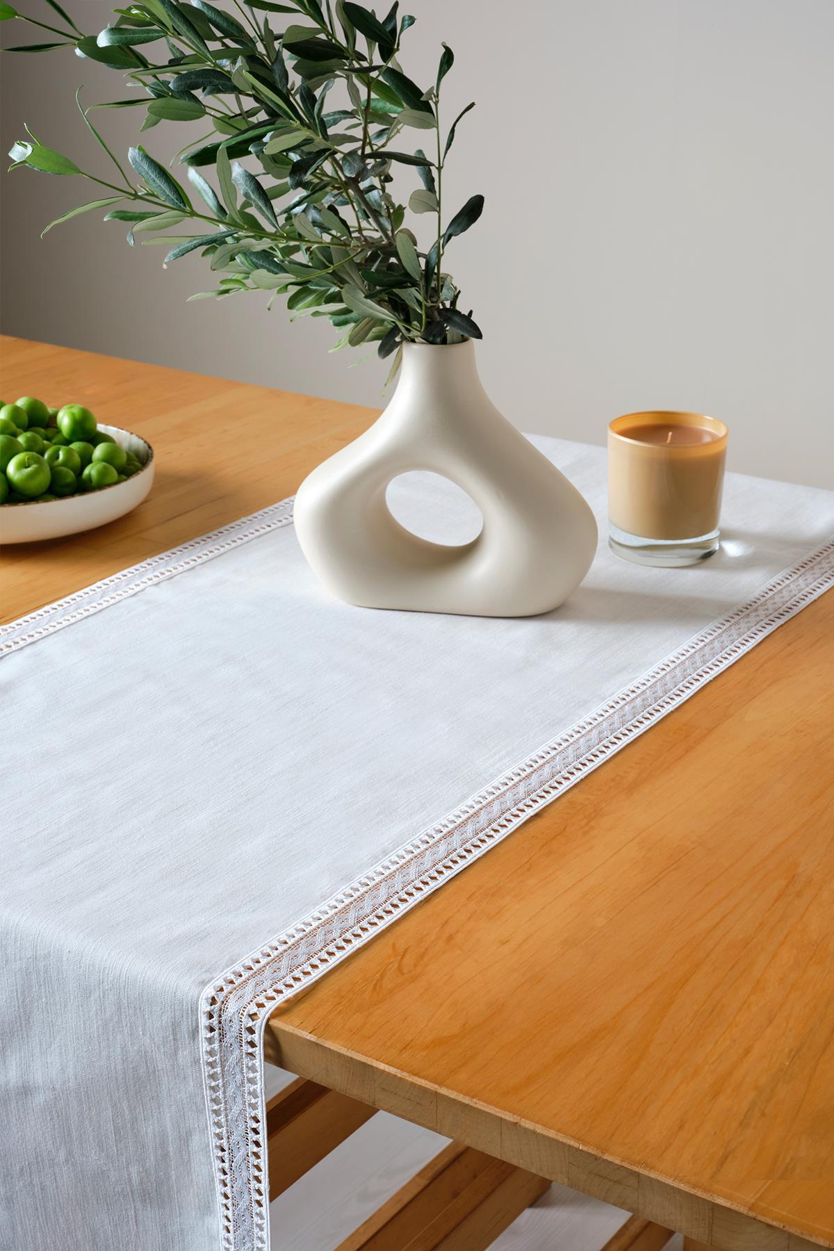 Weißer Spitzen-Tischläufer auf einem rechteckigen Holztisch, dekoriert mit einer modernen Vase, grünen Früchten und einer Kerze.