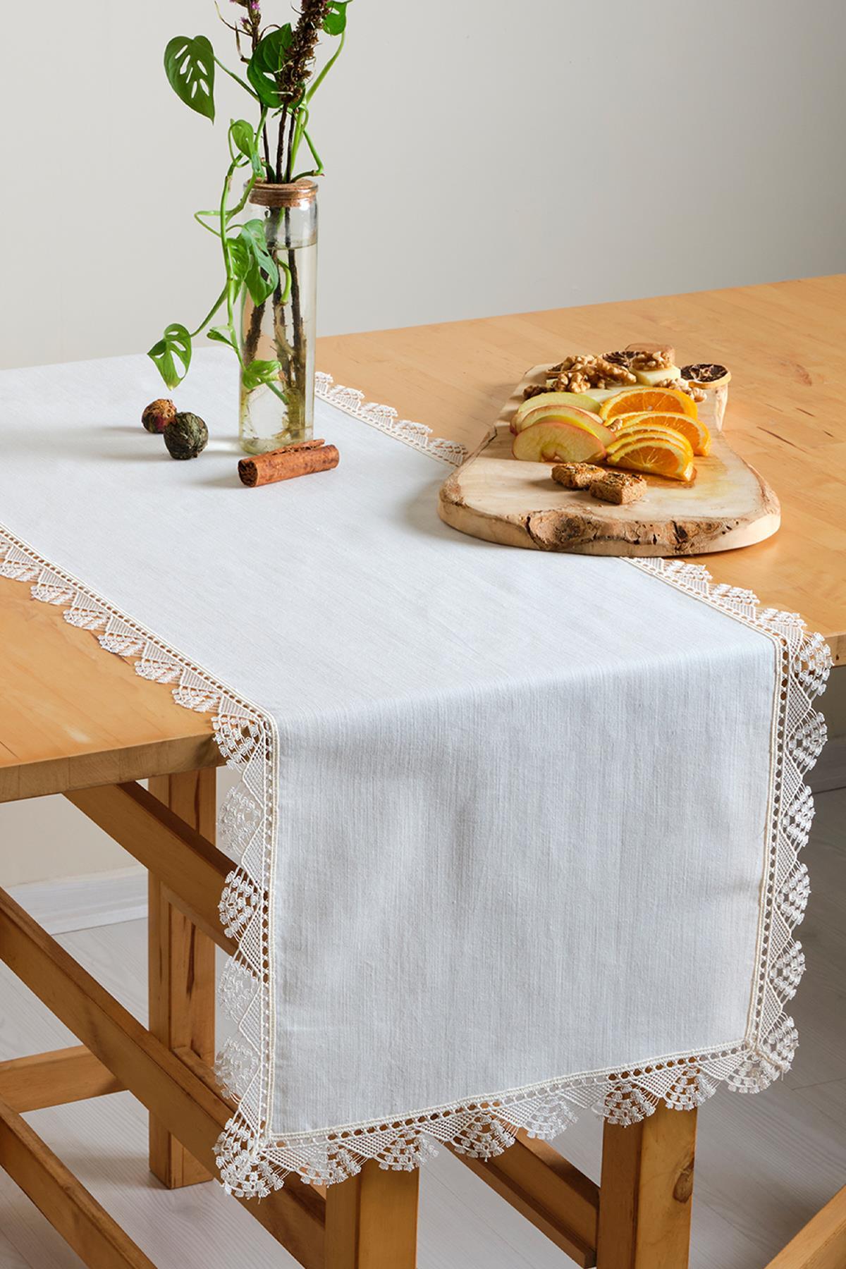 Weißer Tischläufer mit Spitze, dekoriert mit Obst und einer Zierpflanze auf einem Holztisch